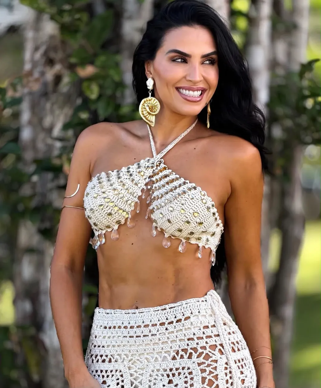 Cliente sorrindo ao ar livre, de top de crochê e brincos dourados, com a pele bronzeada.
