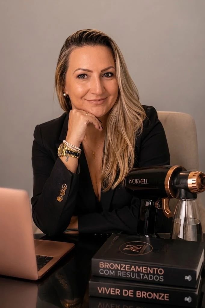 Mariana, da DM Bronzeamento, sorrindo com a mão no queixo, sentada à mesa ao lado do equipamento de aplicação Norvell e de livros sobre bronzeamento.