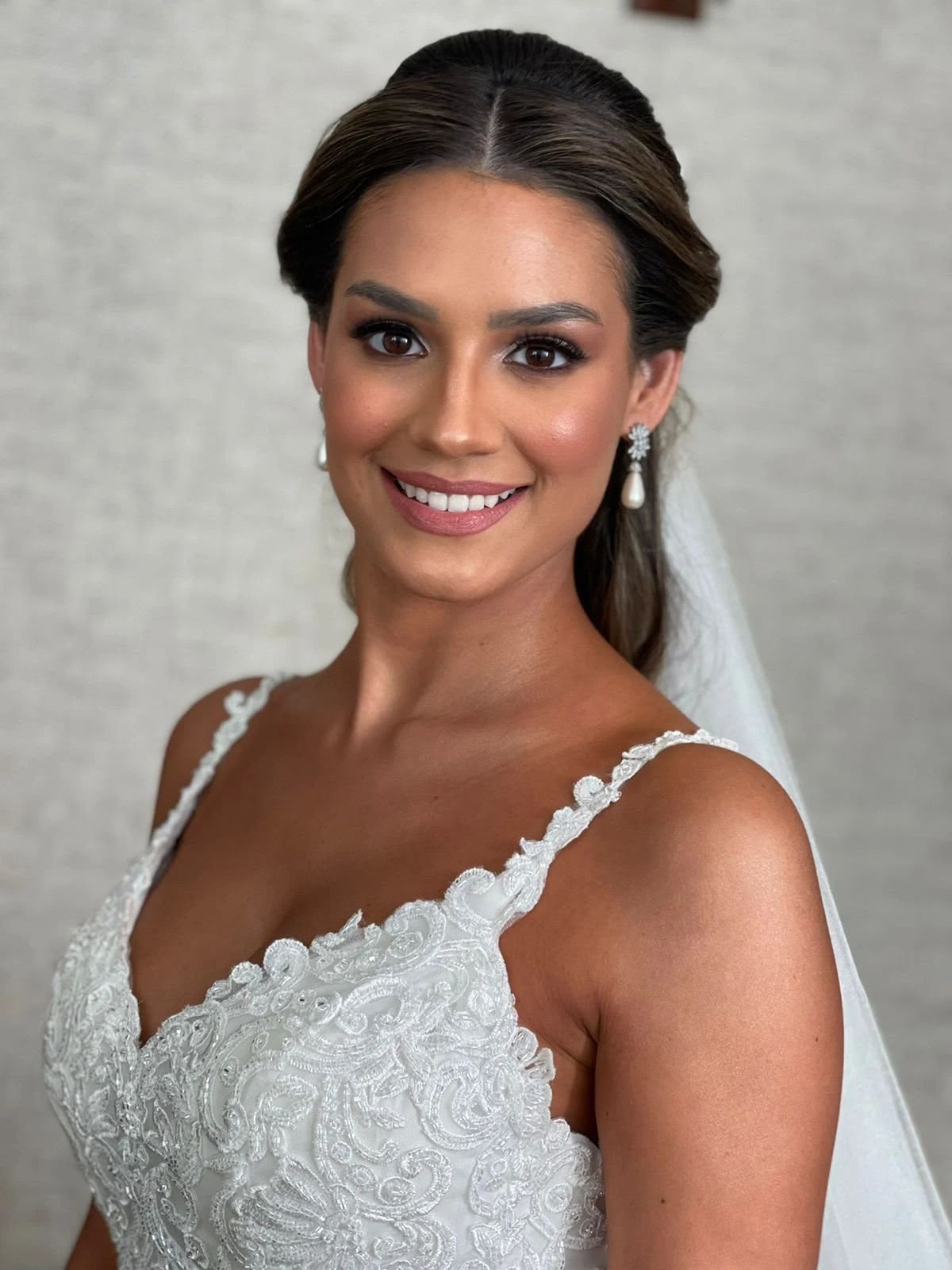 Noiva sorrindo, com vestido de renda, brincos de pérola e véu, a pele com bronze natural.