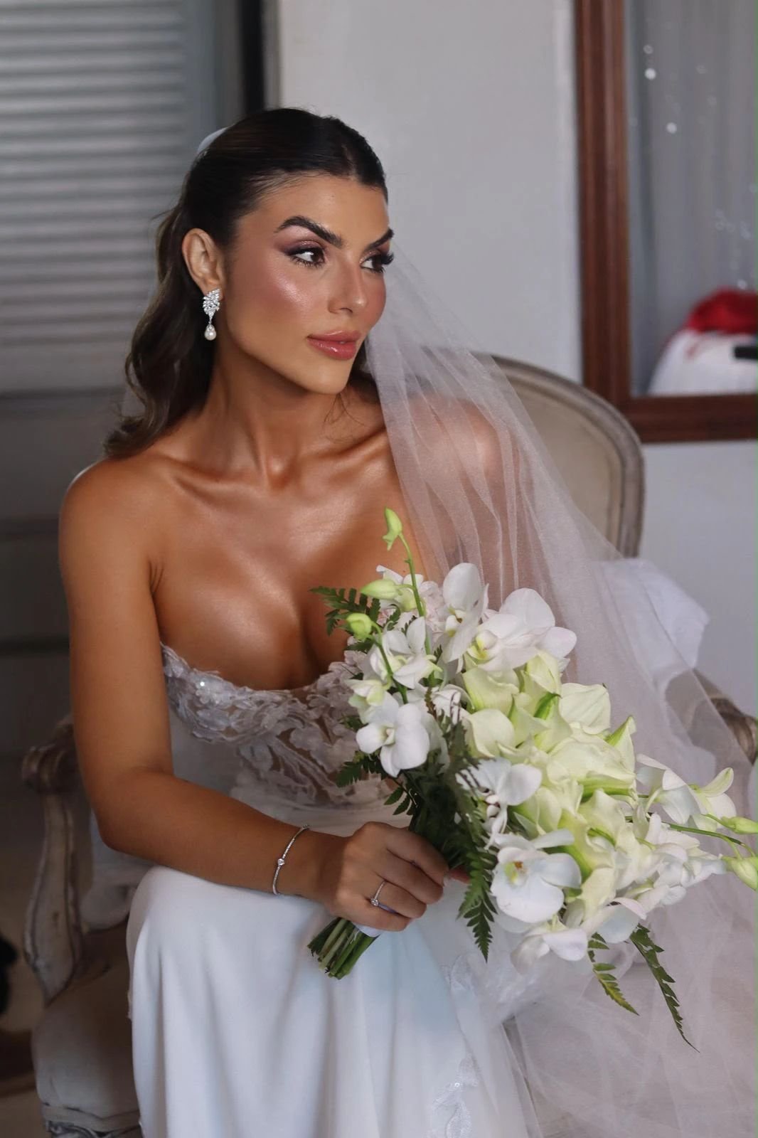 Noiva sentada segurando um buquê de orquídeas brancas, com véu e a pele bronzeada.