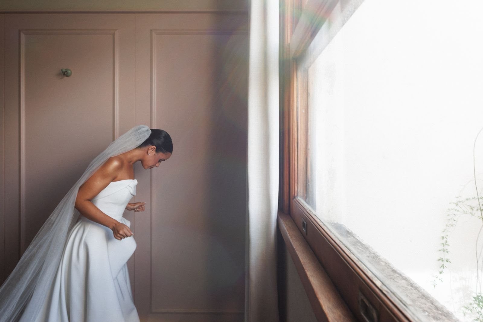 Noiva de véu longo e vestido tomara que caia, inclinada em uma sala de paredes rosadas, perto de uma janela iluminada.