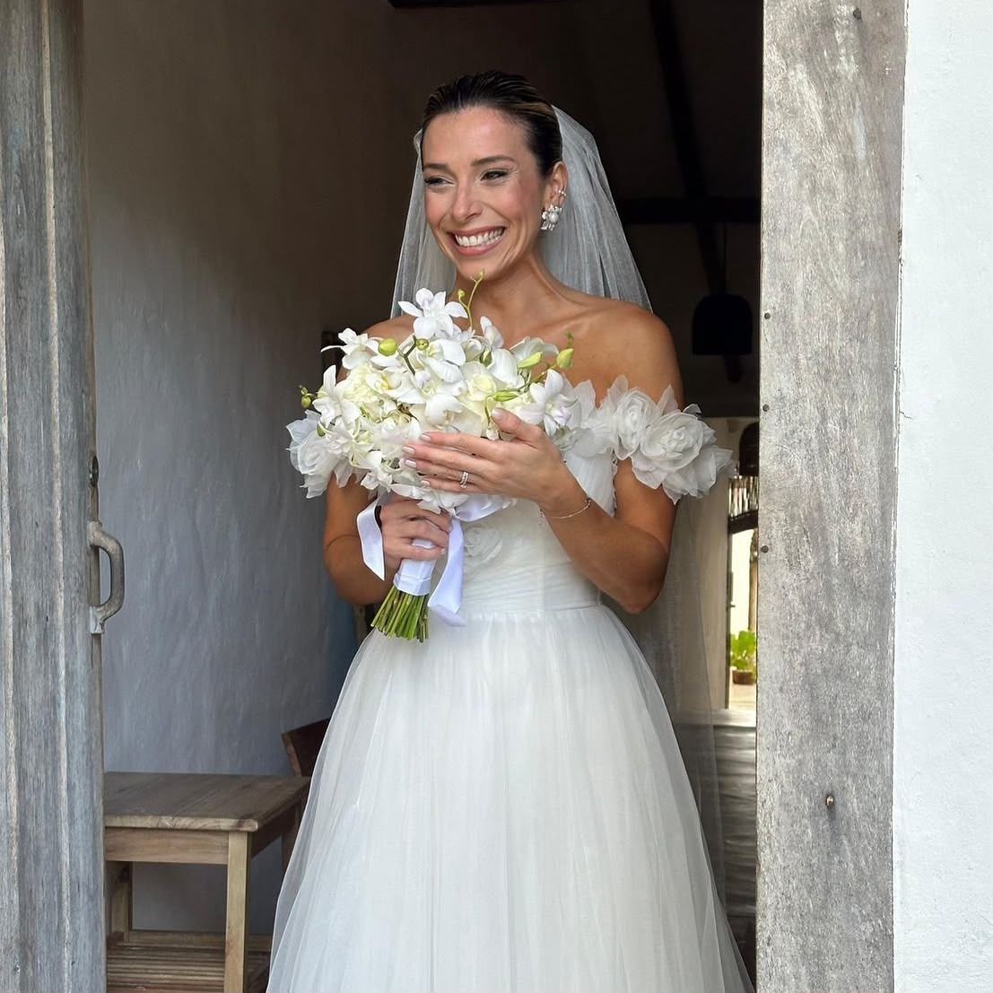 Noiva sorrindo na porta, com buquê de orquídeas brancas, véu e vestido de ombros de fora com flores.